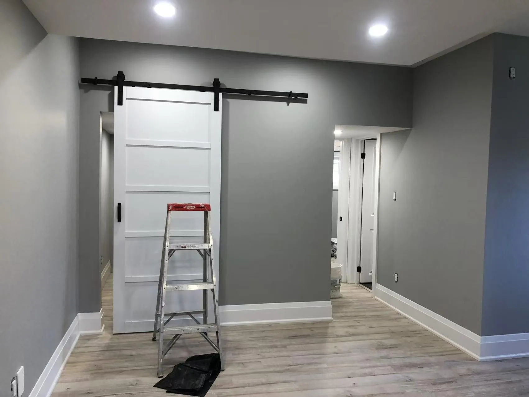 A grey room with a white sliding barn door on black hardware, plank flooring and tall white baseboards.