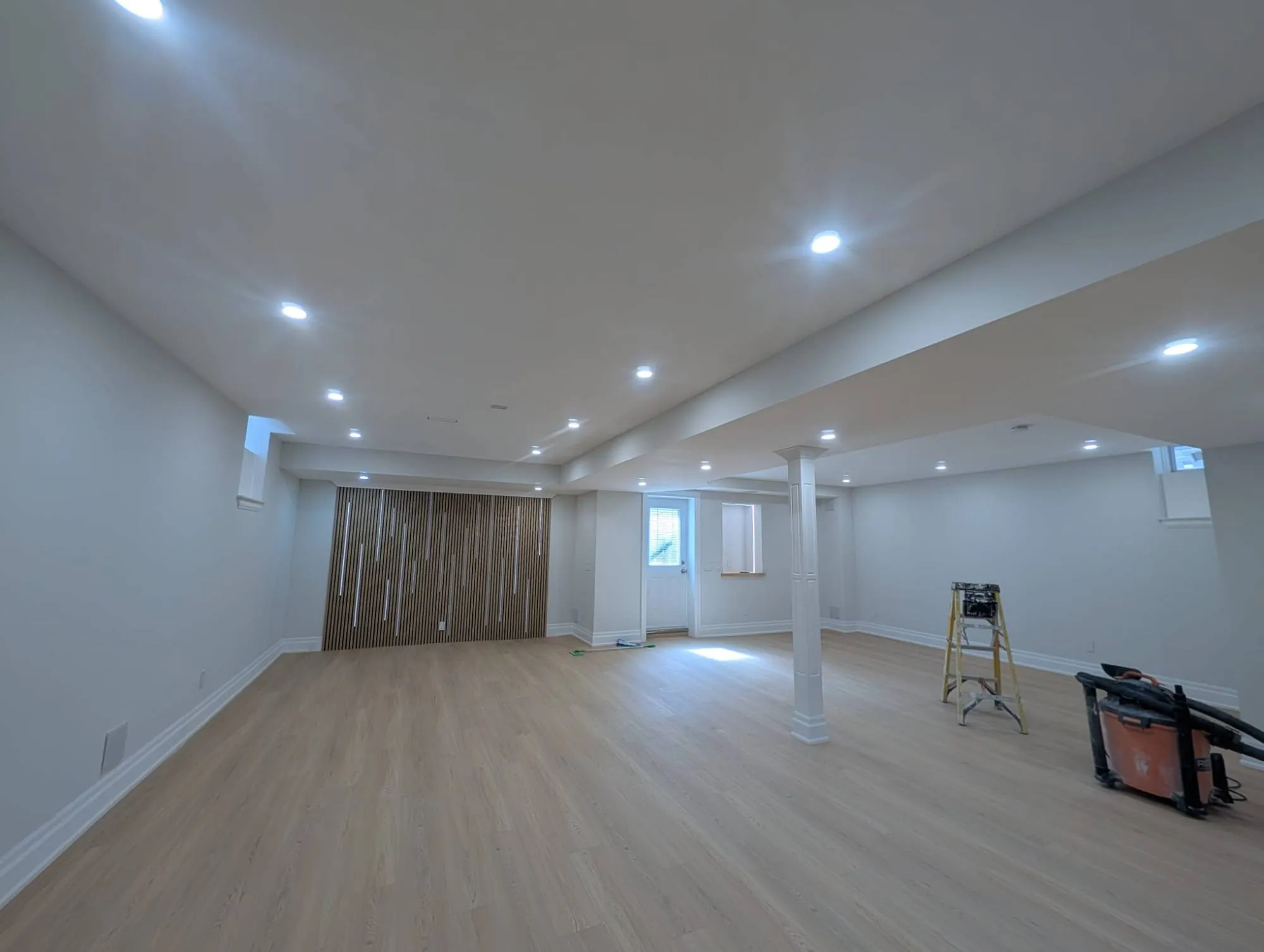 A finished basement with pot lights, wide plank flooring, a wrapped column and a slatted feature wall with lighting.