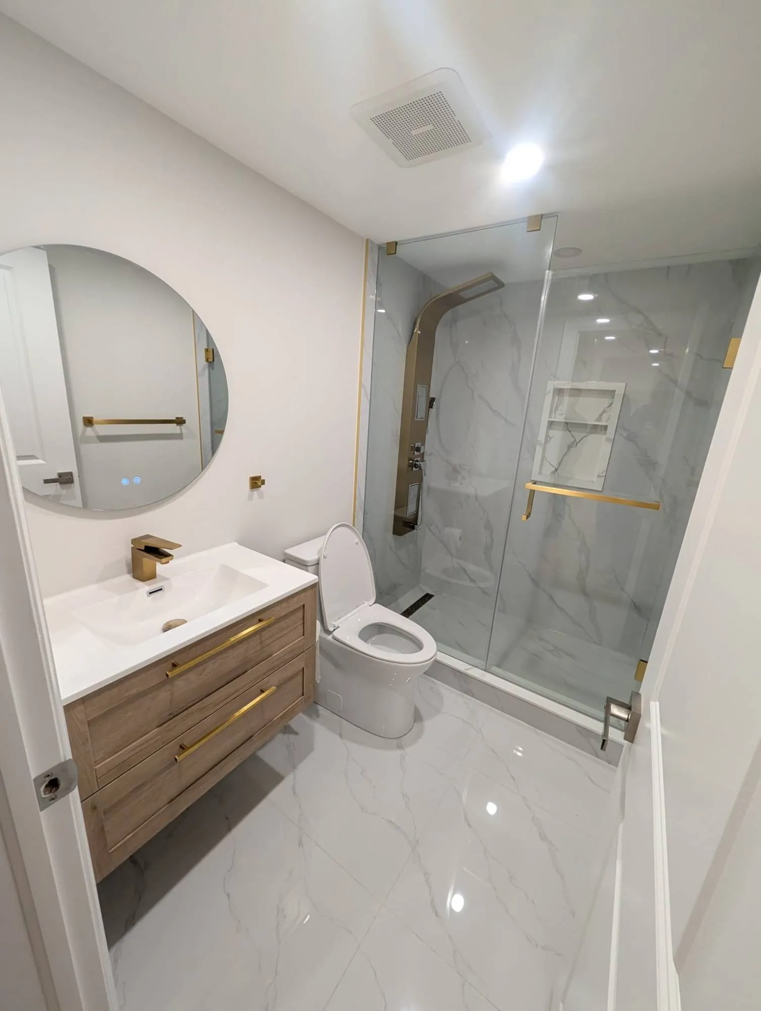 A bathroom in white marble-look tile with brushed gold fixtures, a glass shower with a gold shower panel and a floating wood vanity.