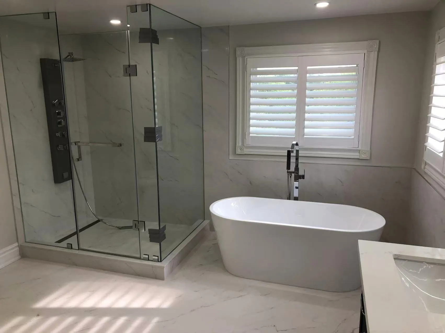 An ensuite with a frameless glass shower, a body-jet shower panel and a freestanding tub under shuttered windows, on large marble-look tile.