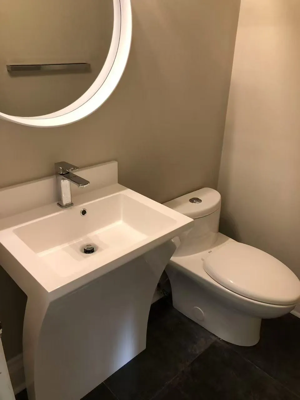 A powder room with a sculpted pedestal sink, a round lit mirror and a one-piece toilet on dark tile.