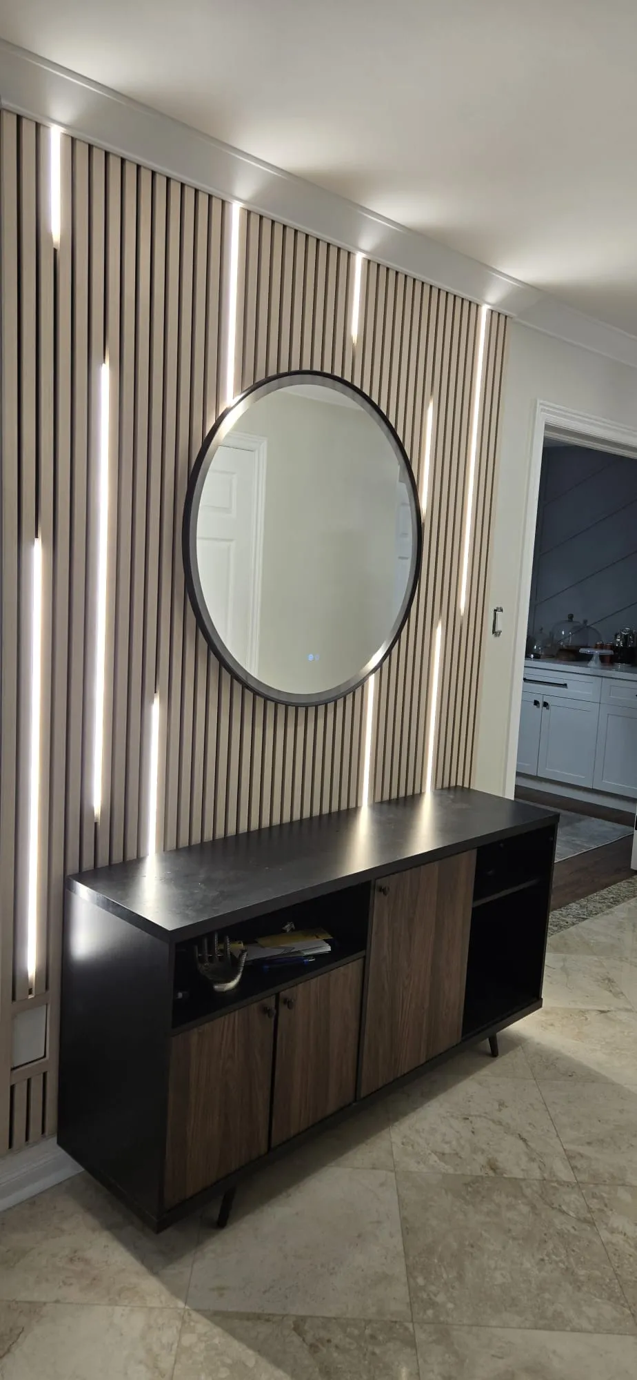The finished slatted wall with built-in light strips and a round mirror above a console.