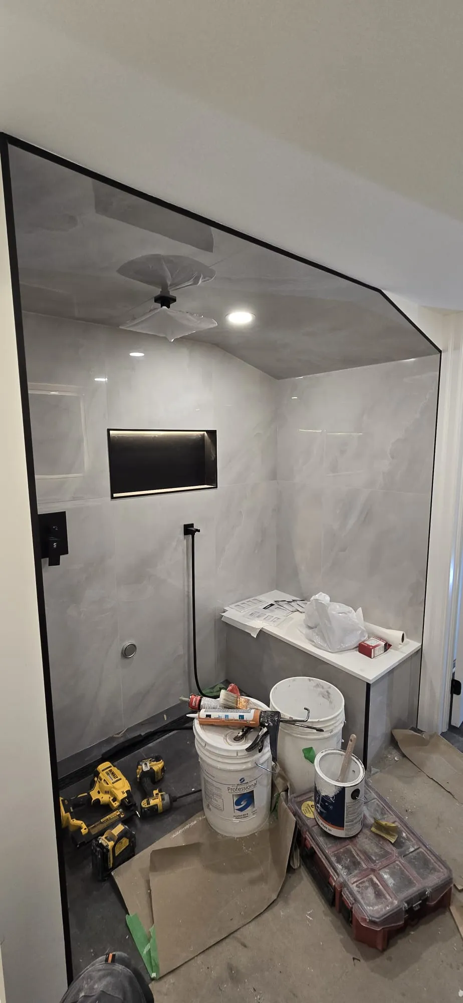 A tiled shower under a sloped ceiling during construction, with a lit black niche, a bench and tools on the floor.