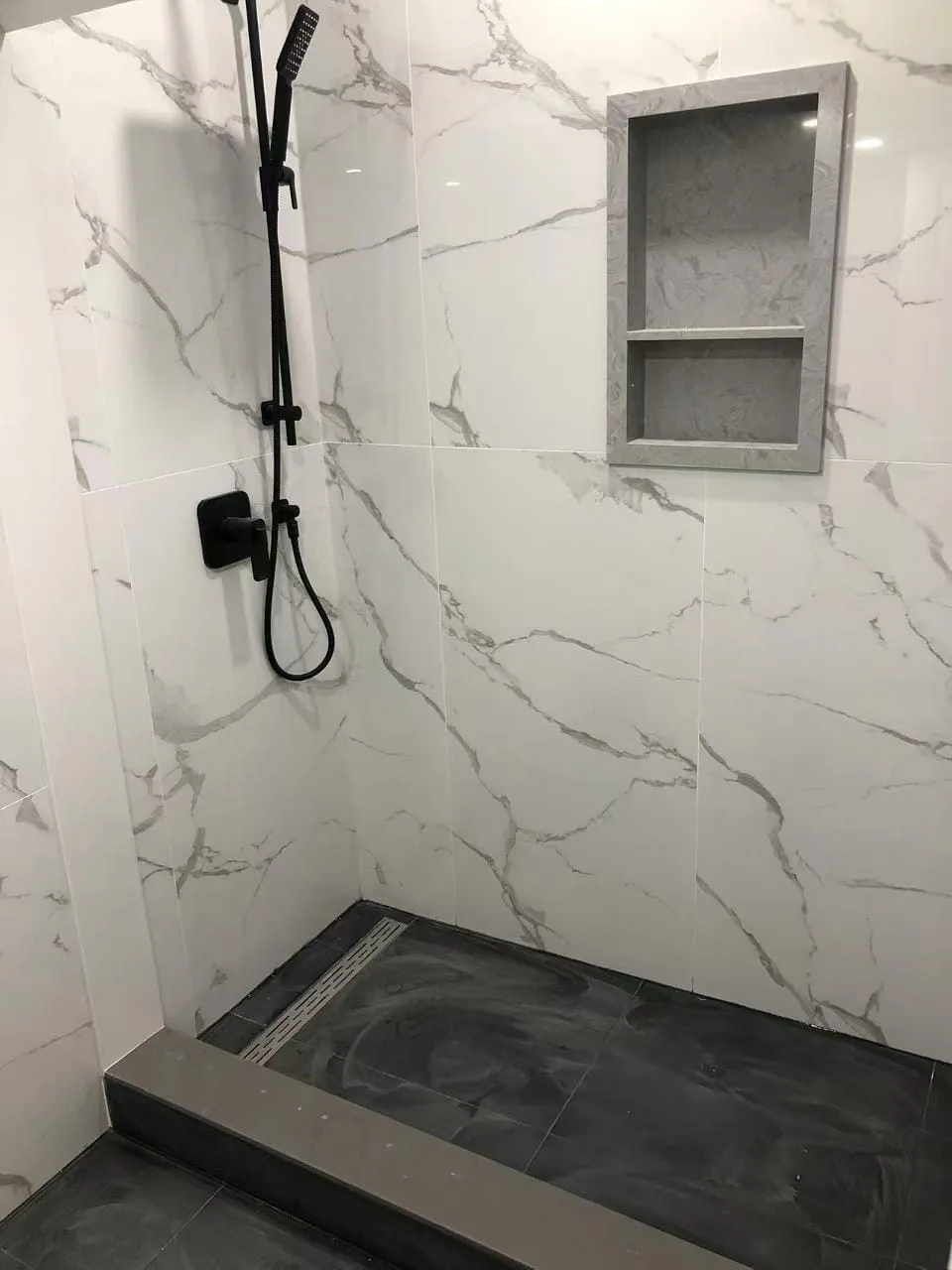 A curbed walk-in shower in marble-look tile with matte black fixtures, a linear drain and a stone niche.